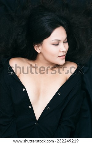 Close up Pretty Young Woman in Unbuttoned Black Blouse, Showing her Cleavage While Lying on Black Cloth Background. Captured in High Angle Shot.