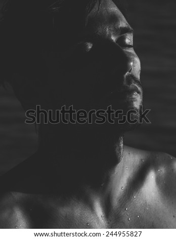 Dark atmospheric portrait of a relaxed young man with a goatee with his head tilted back and eyes closed in meditation, close up head and shoulders with moisture beaded on his skin