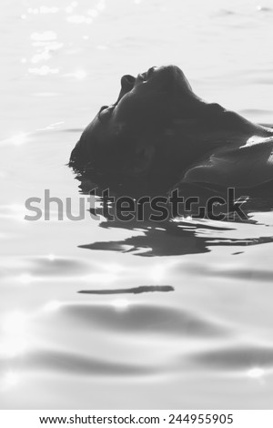 Head of an attractive young woman floating on water with her face up towards the sun, atmospheric greyscale image with rippling water and copyspace
