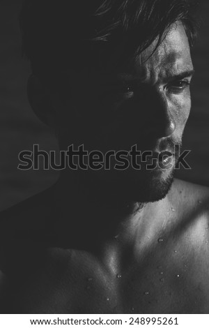 Dark moody portrait of an intense young man with a goatee frowning as he looks to the side, close up of his face and wet bare shoulders