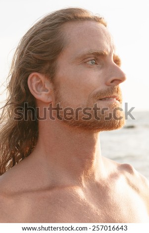 Handsome bearded man with shoulder length hair standing and at the beach staring into the summer sunshine with an intense serious expression and frown