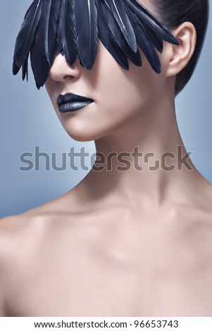 Glamour and beauty shot of a beautiful model with creative make-up of black bird feathers concealing her eyes like a mask. High-end retouching.