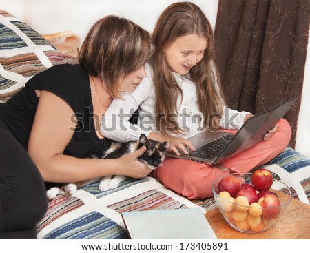 Relaxing family. Caucasian mother and daughter using laptop in the bedroom with smile. Woman and girl are happy. Black cat and vase with peaches next to them. Close up. Indoor.