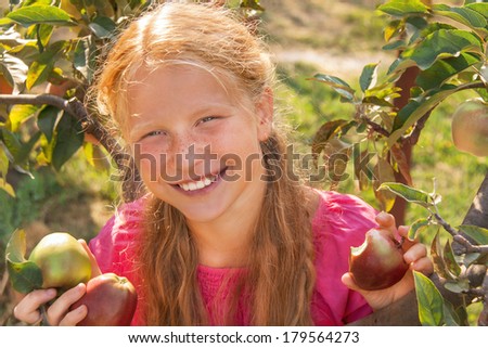 Portrait of happy smiling cute child (girl) with green and red fresh apples near apple tree on beautiful day. Kid has healthy food outside in summer dress and laughing. Outdoor. Close up.
