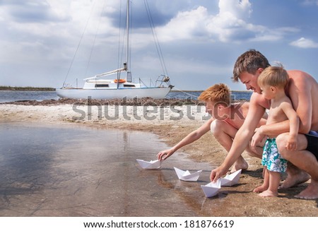 Family vacation. Caucasian happy family (father and sons) is sitting  at the beach near yacht. They are playing with paper boat. Copy space. Outdoor.