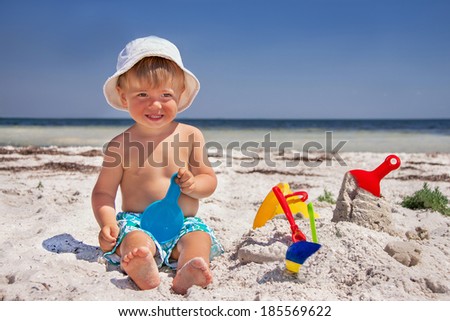 Happy caucasian baby (boy) is sitting and smiling at the beach. Child is playing with toys in summer day.  Travel concept. Outdoor. Close up.