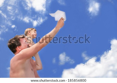 Happy caucasian son and father with paper airplane standing and playing on sky background at the beach. Summer day. Family vacation. Outdoor. Copy space.