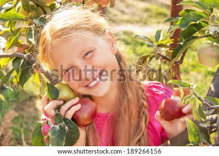 Portrait of happy smiling cute child (girl) with green and red fresh apples near apple tree on beautiful day. Kid eating healthy food outside in summer dress and laughing. Outdoor. Close up.