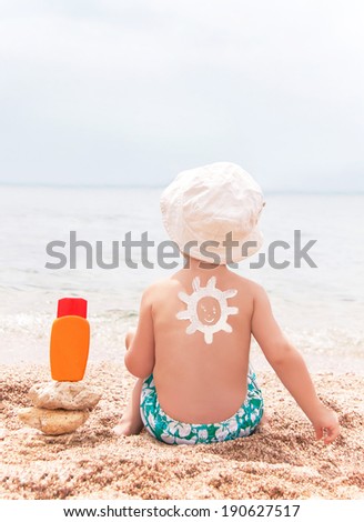 The sun drawing sunscreen (suntan lotion) on baby (boy)  back. Caucasian child is sitting with plastic container of sunscreen on beach. Copyspace. Close up, outdoor (Sharm El Sheikh, Egypt).