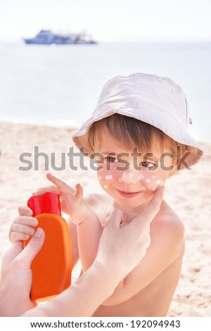 Sun protection. Hand of caucasian mother applying suncream  (suntan lotion) from a plastic container to her happy cute son before tanning during summer holiday on beach. Summer vacations concept.
