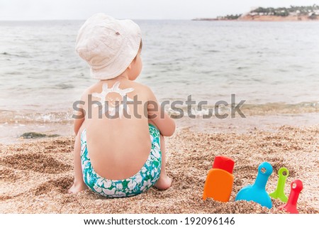 The sun drawing sunscreen (suntan lotion) on baby (boy)  back. Caucasian child is sitting with plastic container of sunscreen and toys on sunny beach. Close up, outdoor (Sharm El Sheikh, Egypt).