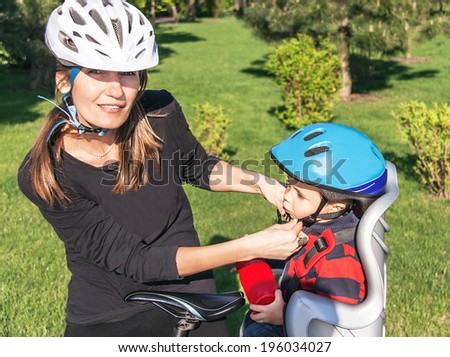 Protection on the bicycle. Caucasian mother have biking helmet and putting biking helmet on outside to her son. The son is in the bicycle chair (seat) during bicycle ride. Travel concept.