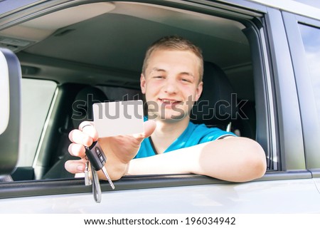 Car driver. Caucasian teen boy showing an empty white card for your message, new car key and car. Happy smiling young man behind the wheel. Travel and rental concept. Copy space, close up.