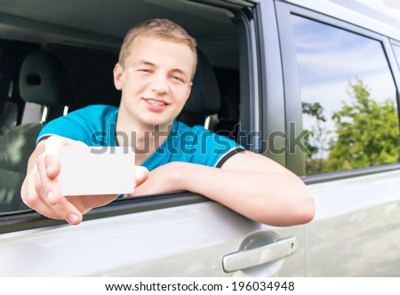 Car driver. Caucasian teen boy showing an empty white card for your message in the new car. Happy smiling young man behind the wheel. Travel and rental concept. Close up, outdoor.