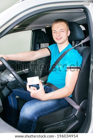 Car driver. Caucasian teen boy fastening seat belt and showing driver license in the car. Happy smiling young man behind the wheel. Travel and rental concept. Close up, outdoor.