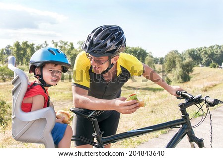 Happy father and son is eating lunch (snack) during bicycle ride. Child (boy) and man have biking helmets. The son is in the bicycle chair (seat). Caucasian smiling male models. Travel concept.