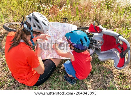 Happy bikers child (boy) and mother is holding a map during bicycle ride. Kid and woman have biking helmets. Toddler has bicycle chair (seat). Caucasian male and female models. Travel concept.