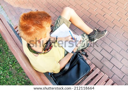High angle portrait of a left-handed teen boy (schoolboy). Teenager is sitting and writing on the bench. Young red-head boy is studying. Caucasian male model (14 year). Sunny day. Education concept.