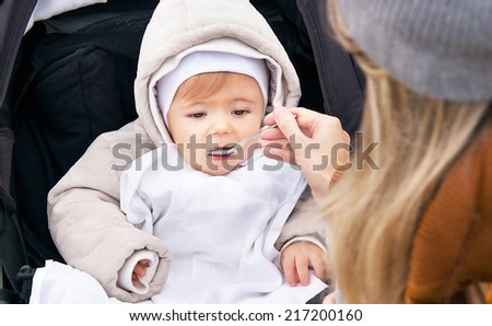 Mother is feeding nine month baby boy from a spoon. Child has healthy food outdoor. Porridge is for healthy eating child (kid). Caucasian male model is sitting in the baby buggy (pram, pushchair).