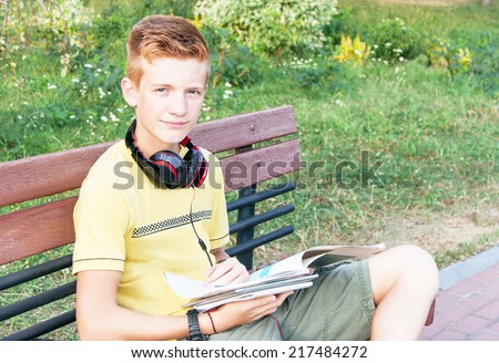 Schoolboy teen (teenager) is sitting with books, head phone on the bench in the city park. Young red-head boy is studying. Caucasian cute male model (14 year). Sunny day. Education concept. Close up.