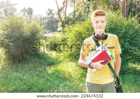 Schoolboy teen (teenager) is standing with books, head phone and school bag in the park. Young child is in the sunny day. Caucasian cute male model (14 year). Education concept. Copy space, outdoors.