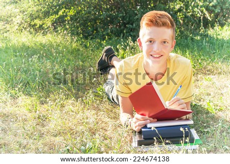 Schoolboy left-handed teen (teenager) lying and writing in the park. Young smiling red-head boy studying on the grass. Caucasian cute male model (14 year). Sunny day. Education concept. Copy space.