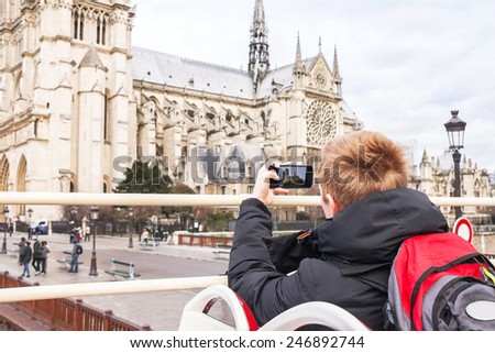 Teenager tourist is taking photo on cathedral of Notre Dame de Paris with mobile smart phone. Left-handed boy is on tourist bus. Caucasian male model. Travel, tourism, vacation concept. France.