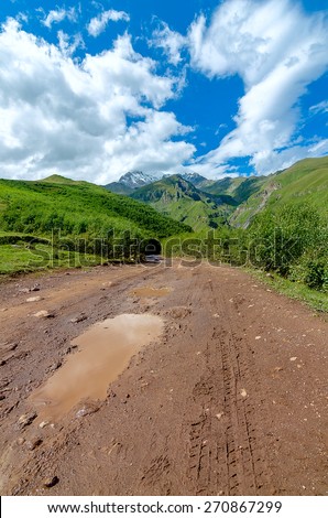 Road with tire tracks leading to mountains and beautiful clouds. Puddle with small stones on the way