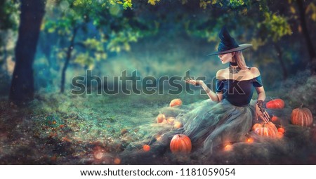 Halloween Witch with a carved Pumpkin and magic lights in a dark forest at night. Beautiful young woman in witches costume pointing hand, holding some product, advertising. Halloween party art design