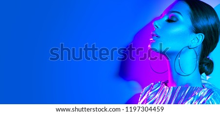 High Fashion model girl in colourful bright neon uv lights posing in studio, portrait of beautiful woman, trendy glowing metallic make-up, silver accessories earrings. Vivid neon makeup. Wide angle
