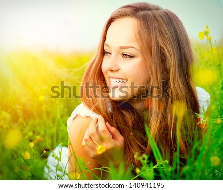 Beauty Girl in the Meadow. Beautiful Young Woman Outdoors. Enjoy Nature. Healthy Smiling Girl lying on Green Grass with wild Flowers. Laughing And Happy