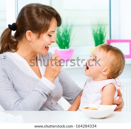 Mother Feeding Her Baby Girl With A Spoon. Mother Giving Food To Her Adorable Child At Home