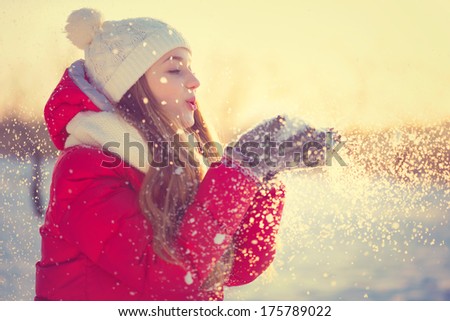 Beauty Winter Girl Blowing Snow in frosty winter Park. Outdoors. Flying Snowflakes. Sunny day. Backlit. Joyful Beauty young woman Having Fun in Winter Park.