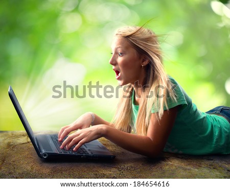 Surprised Teenage Girl with Laptop outdoors. Young woman using pc outdoor laying on grass. Nature background.