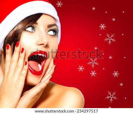 Christmas Surprised Winter Woman. Beauty Model Girl in Santa Hat over holiday red Background. Funny Laughing Woman Portrait. Open Mouth. True Emotions. Red Lips and Manicure. Beautiful Holiday Makeup