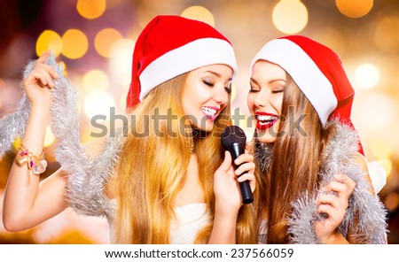 Christmas party, karaoke. Beauty girls in santa hat with a microphone singing and dancing over holiday glowing background. Disco. New year celebration