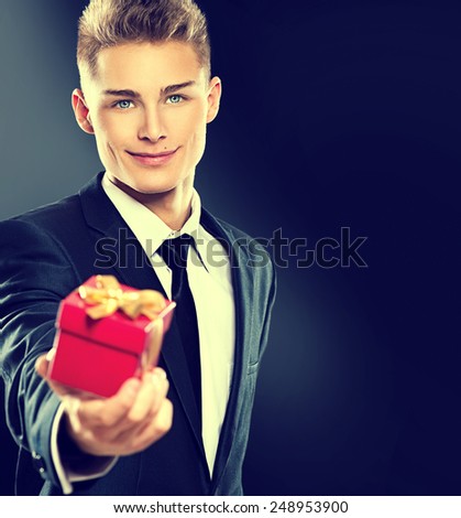 Young handsome man giving red gift box. Elegant businessman in dark suit over black background. Sexy blonde smiling guy with holiday Valentine\'s Day gift