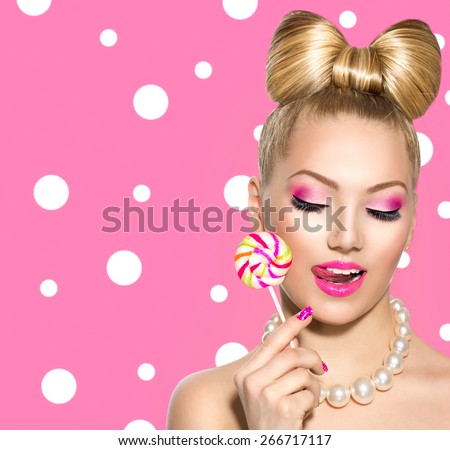 Beauty fashion model girl Eating colourful lollipop. Young funny woman with bow hairstyle, pink nail art and makeup over polka dots background. Sweet candy