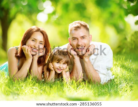 Happy joyful young family father, mother and little daughter having fun outdoors, playing together in summer park. Mom, Dad and kid laughing, lyying on green grass, enjoying nature outside. Sunny day