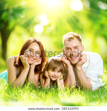 Happy joyful young family father, mother and little daughter having fun outdoors, playing together in summer park. Mom, Dad and kid laughing, lyying on green grass, enjoying nature outside. Sunny day