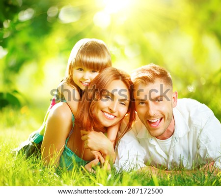 Happy joyful young family father, mother and little daughter having fun outdoors, playing together in summer park. Mom, Dad and kid laughing, lying on green grass, enjoying nature outside. Sunny day