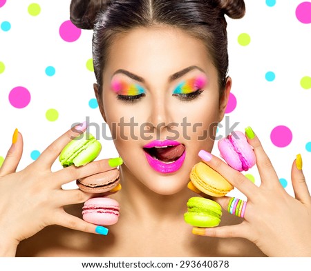 Beauty fashion model girl with colourful makeup and manicure taking colorful macaroons. Beautiful woman, bright make-up. Purple lipstick, vivid eyeshadow and accessories. Diet, dieting concept. Sweets