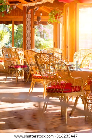 Restaurant interior. Fashioned cafe terrace. View of an sunny empty summer coffee terrace with tables and wicker chairs. Interior of cozy restaurant decorated with plant. Vertical photo
