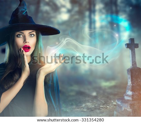 Halloween Witch with a magic in a dark Old Spooky cemetery. Beautiful young woman in witches hat and costume holding magical light in her hand. Halloween art design