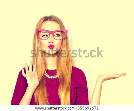 Beauty funny teenage girl with paper glasses on stick showing empty copy space on the open hand palm for text. Happy girl presenting point, looking at camera. Proposing product. Advertisement gesture