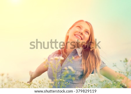 Beauty Girl Outdoors enjoying nature. Beautiful Teenage Model girl having fun on spring Field with blooming flowers, raising hands in Sun Light. Sunrise. Glow Sun. Free Happy Woman, allergy free