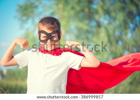 Super Hero Kid having fun outdoor. Superhero little boy over nature green blurred background showing muscles. Little boy wearing superhero costume and having fun outdoors
