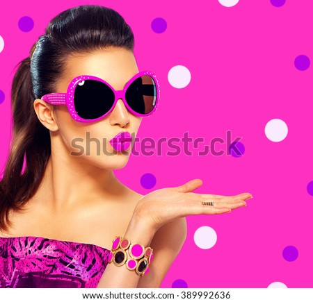 Beauty surprised fashion model girl wearing purple sunglasses, with bright makeup showing empty copy space on open hand palm for text over pink background. Emotions. Beautiful woman, summer fashion