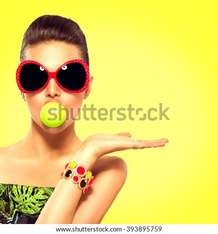 Beauty summer fashion model girl wearing sunglasses with green bubble of chewing gum and bright makeup showing empty copy space on the open hand palm for text over yellow background. Beautiful woman