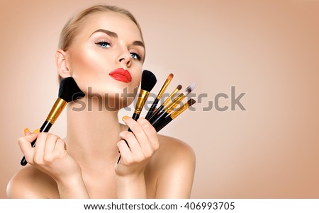 Beauty Woman with Makeup Brushes. Natural Make-up for Blonde Model Girl with Blue Eyes. Beautiful Face. Perfect Skin. Applying Holiday Makeup, orange color lipstick and manicure over beige background